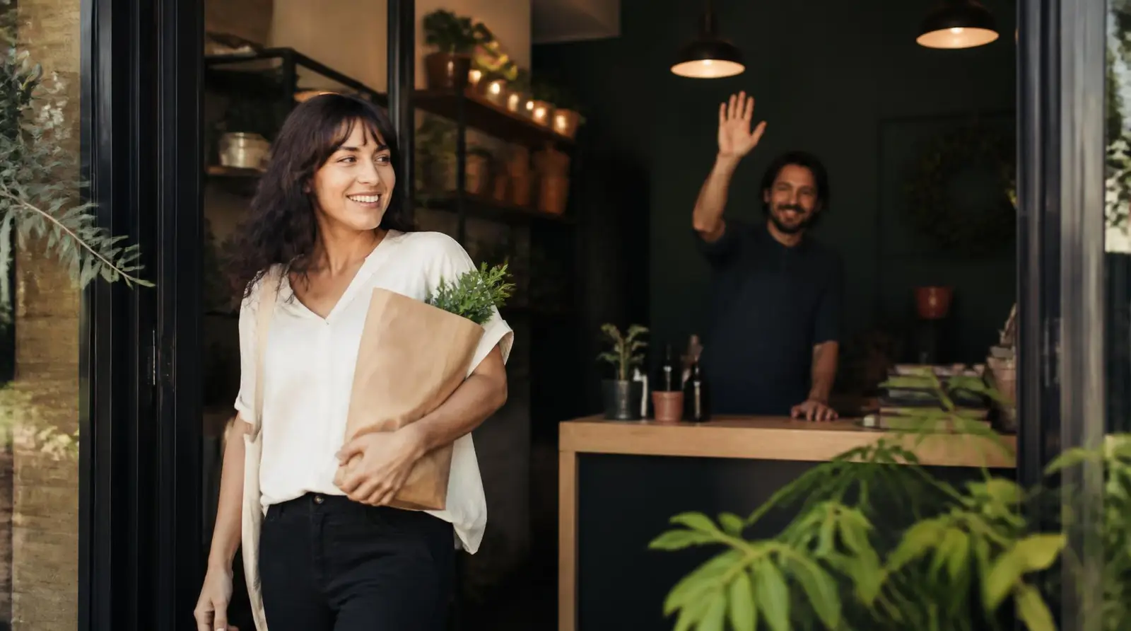 Customer leaving a small shop with a paper bag while the owner waves goodbye
